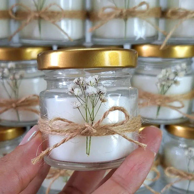Hand holding a Fiorance Luxe luxury soy wax candle in a glass jar with gold lid, twine bow, and small white flowers, with similar candles in the background.