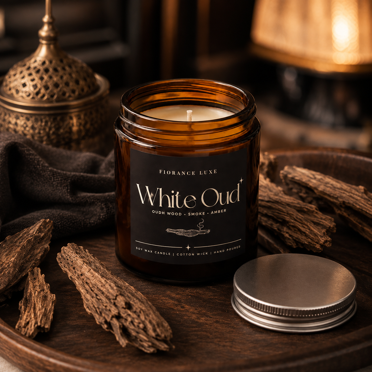 An amber glass jar candle labeled 'White Oud' by Flandrance Luxe, shown on a dark wooden tray with dried botanicals and a silver metal lid beside it. The background includes warm ambient lighting and decorative objects.