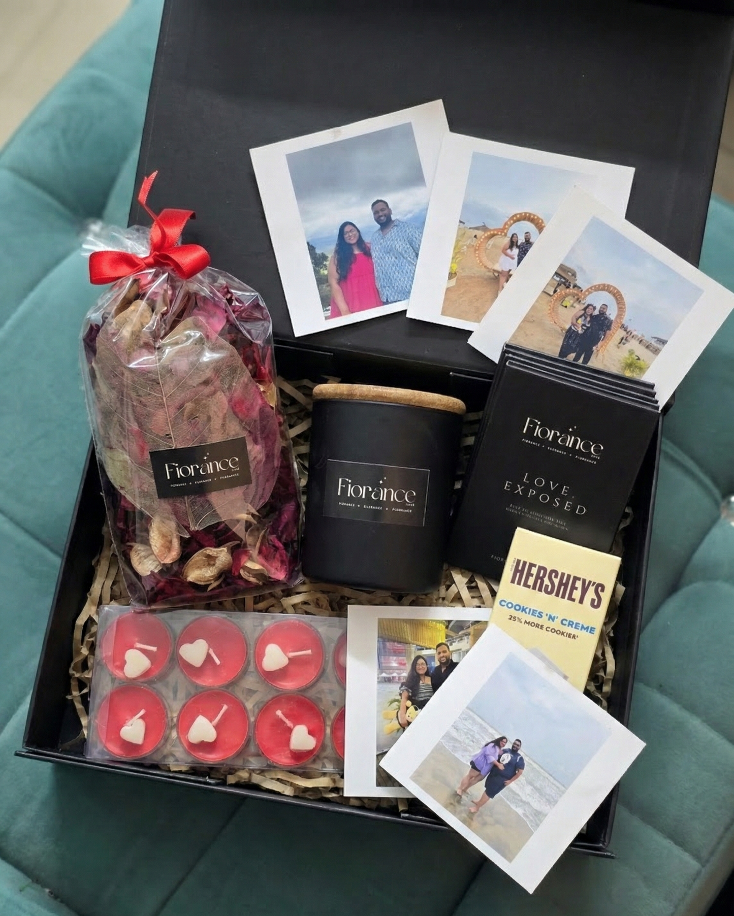 Gift box with assorted items including a candle, cookies, and photos on a teal surface.