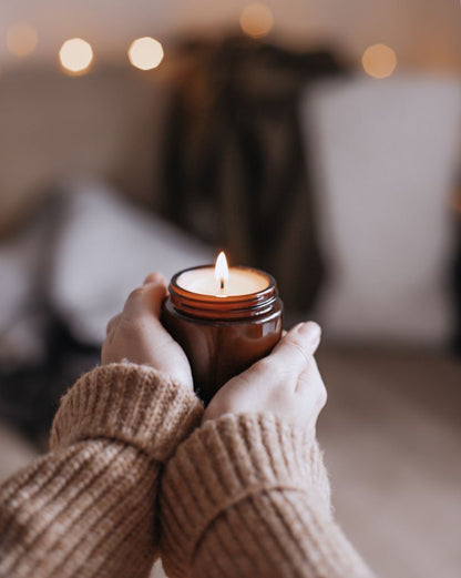 Hands holding a lit soy wax scented candle in a brown jar, cozy home decor setting