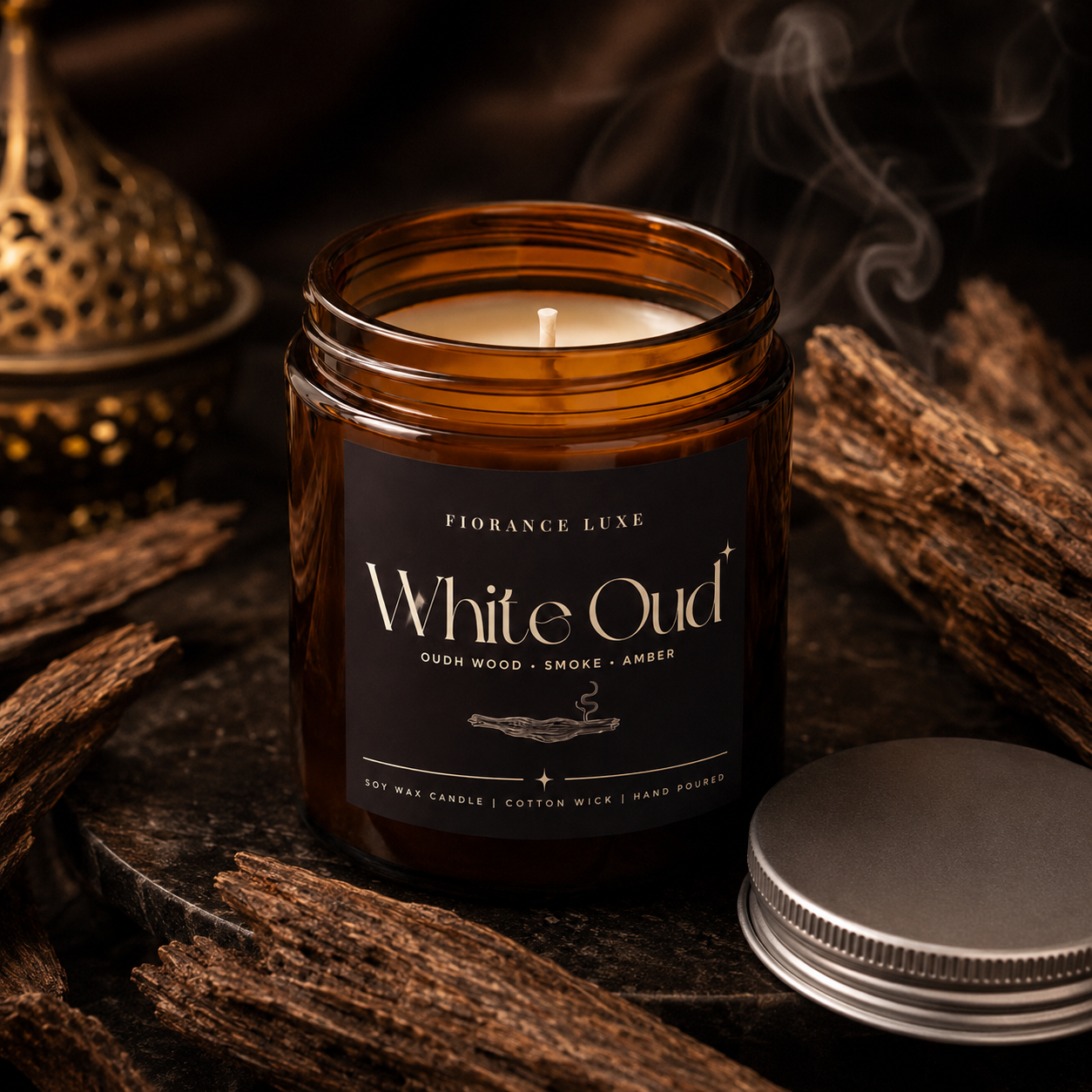 An amber glass jar candle labeled 'White Oud' by Fiorance Luxe, shown with its silver lid beside it on a dark rustic surface surrounded by wood pieces and smoke.