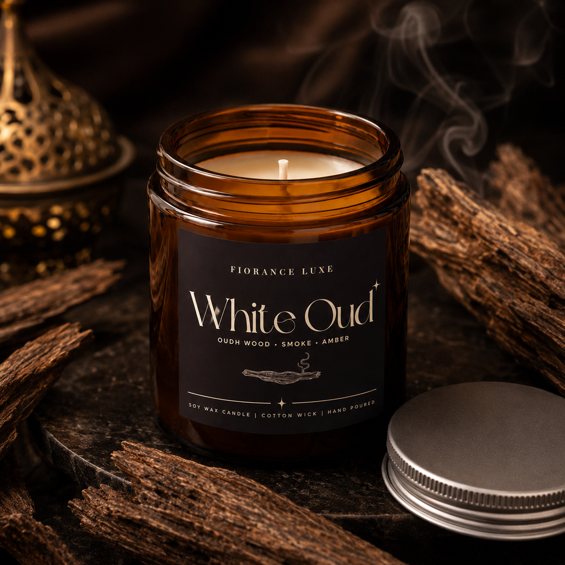 An amber glass jar candle labeled 'White Oud' by Fiorance Luxe, shown with its silver lid beside it on a dark rustic surface surrounded by wood pieces and smoke.